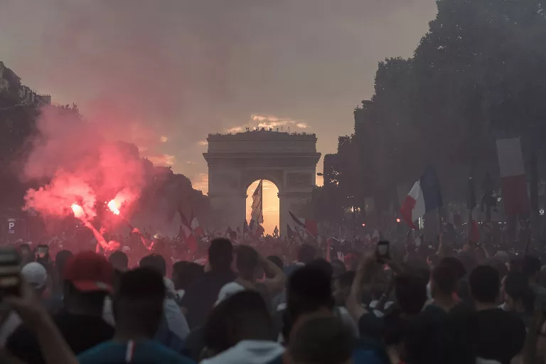 Une foule dense envahit les Champs-Élysées au crépuscule, agitant des drapeaux tricolores et allumant des fumigènes rouges, l'Arc de Triomphe se découpant sur un coucher de soleil orangé.